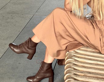 May include: A pair of dark brown leather ankle boots featuring a lace-up front closure, a rounded toe, and a block heel, paired with a long, solid tan-colored dress made of a smooth, slightly reflective fabric that falls to the mid-calf.