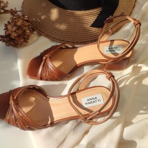 Puede incluir: Sandalias de cuero marr&oacute;n con correas en el tobillo y tac&oacute;n bajo. Las sandalias presentan un dise&ntilde;o anudado en la correa del dedo y la marca "ANNA MARATTI" es visible. Un sombrero de paja est&aacute; en el fondo. Altura del tac&oacute;n: unos 5 cm.