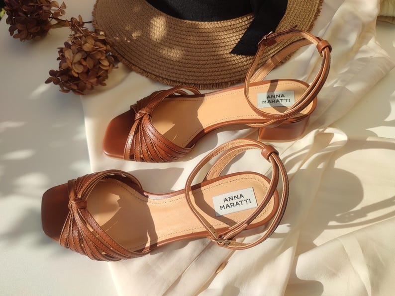 Puede incluir: Sandalias de cuero marr&oacute;n con correas en el tobillo y tac&oacute;n bajo. Las sandalias presentan un dise&ntilde;o anudado en la correa del dedo y la marca "ANNA MARATTI" es visible. Un sombrero de paja est&aacute; en el fondo. Altura del tac&oacute;n: unos 5 cm.