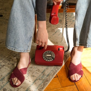 Pode incluir: Um telefone vermelho vintage com disco rotativo, com um auscultador e cabo a condizer. O telefone est&aacute; sobre um tapete estampado, perto de um par de sand&aacute;lias bord&ocirc; com tiras cruzadas. As sand&aacute;lias s&atilde;o usadas com jeans azul claro.