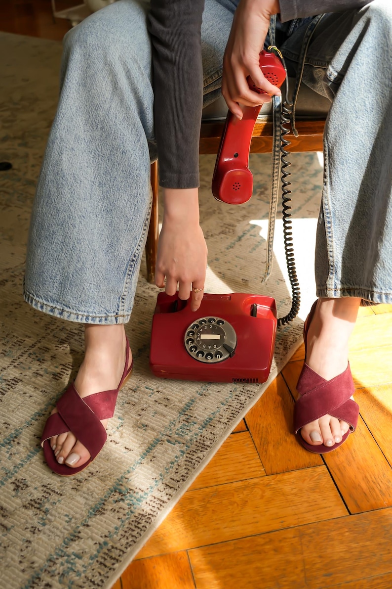 Pode incluir: Um telefone vermelho vintage com disco rotativo, com um auscultador e cabo a condizer. O telefone est&aacute; sobre um tapete estampado, perto de um par de sand&aacute;lias bord&ocirc; com tiras cruzadas. As sand&aacute;lias s&atilde;o usadas com jeans azul claro.