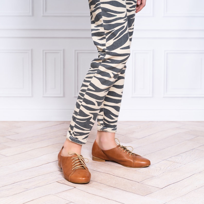 May include: A pair of brown leather lace-up shoes with a low heel. The shoes are worn by a person wearing zebra print pants.