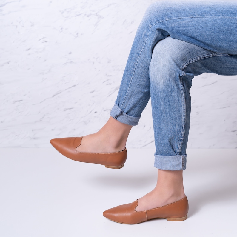 Op de afbeelding: Een paar bruine leren spitse ballerina's. De schoenen worden gedragen door iemand die een blauwe jeans draagt.