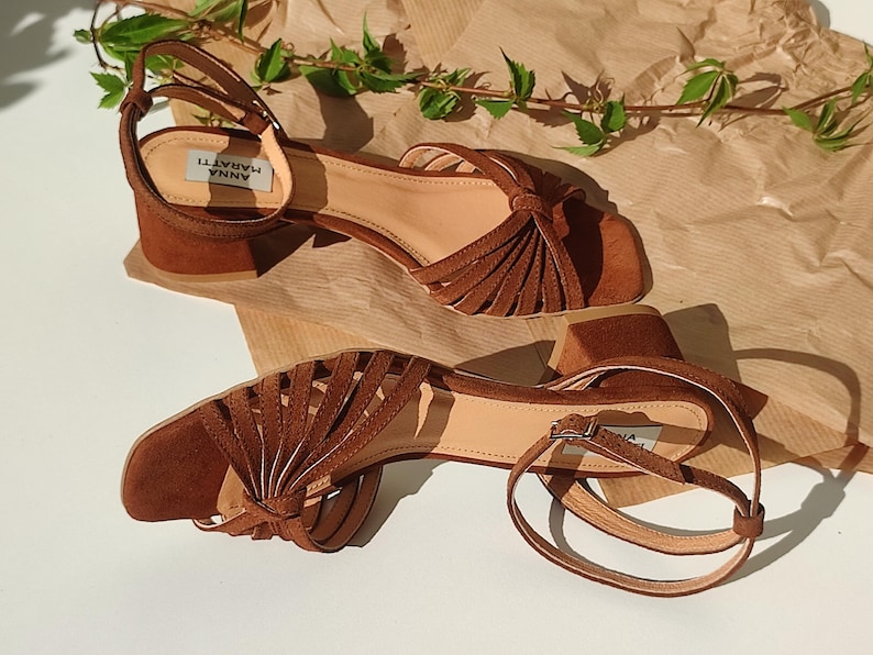 Puede incluir: Sandalias de ante marr&oacute;n con tac&oacute;n ancho. Las sandalias presentan punta cuadrada, correa en el tobillo y detalles de flecos decorativos. La marca "ANNA MARATTI" es visible en la plantilla. Fondo neutro.