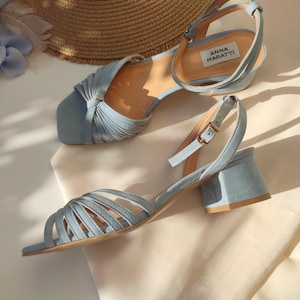 Może przedstawiać: Jasnoniebieskie sandały na słupku z paskiem na kostce. Sandały mają kwadratowy nosek i wiele pask&oacute;w na wierzchu. Marka "ANNA MARATTI" jest widoczna na wkładce. Słomkowy kapelusz i niebieskie kwiatowe akcenty w tle.