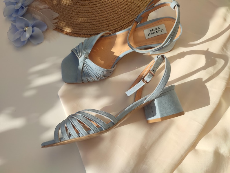 Może przedstawiać: Jasnoniebieskie sandały na słupku z paskiem na kostce. Sandały mają kwadratowy nosek i wiele pask&oacute;w na wierzchu. Marka "ANNA MARATTI" jest widoczna na wkładce. Słomkowy kapelusz i niebieskie kwiatowe akcenty w tle.