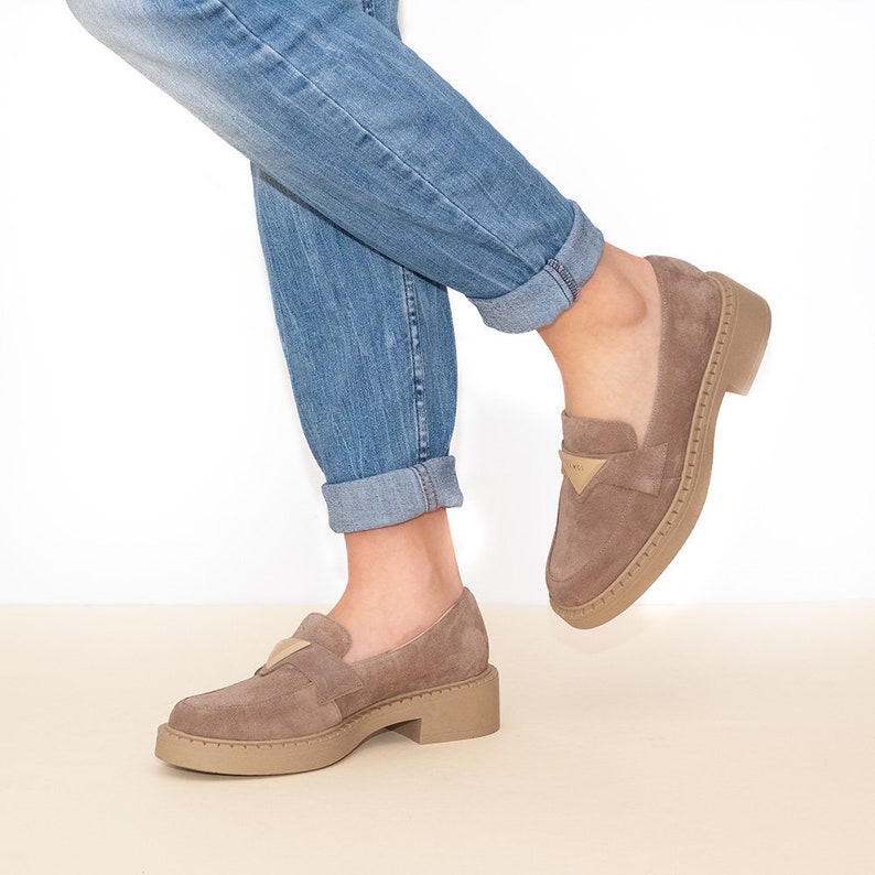 May include: A pair of light brown suede loafers with a chunky platform sole. The loafers have a gold accent on the front and are worn with rolled up blue jeans.