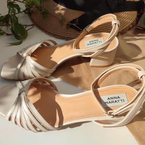 May include: A pair of cream-colored sandals with a square toe and ankle strap. The sandals have a low block heel and feature a woven design across the top. The brand name "ANNA MARATTI" is visible on the insole.