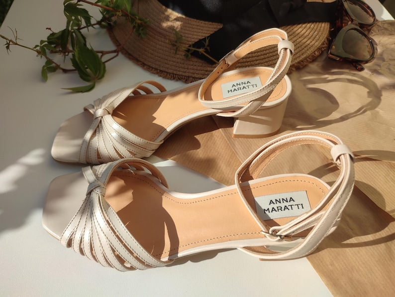May include: A pair of cream-colored sandals with a square toe and ankle strap. The sandals have a low block heel and feature a woven design across the top. The brand name "ANNA MARATTI" is visible on the insole.