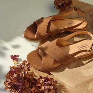 Pode incluir: Um par de sand&aacute;lias de camur&ccedil;a castanhas com tiras cruzadas e tiras no tornozelo. As sand&aacute;lias t&ecirc;m um salto pequeno e um fecho de fivela. A marca "Anna Maratti" &eacute; vis&iacute;vel na palmilha. As sand&aacute;lias est&atilde;o numa superf&iacute;cie castanha clara.