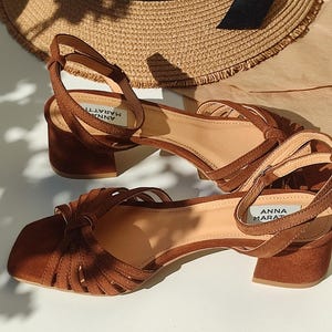 Puede incluir: Sandalias de ante marr&oacute;n con punta cuadrada y tac&oacute;n ancho. Las sandalias tienen m&uacute;ltiples correas en la parte superior y una correa en el tobillo con el nombre de la marca "ANNA MARATTI". Un sombrero de paja en el fondo.