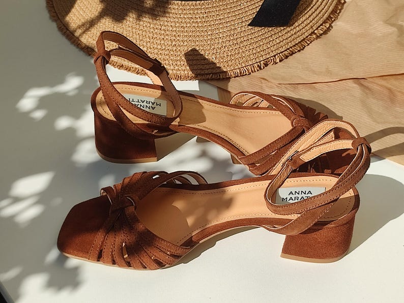 Puede incluir: Sandalias de ante marr&oacute;n con punta cuadrada y tac&oacute;n ancho. Las sandalias tienen m&uacute;ltiples correas en la parte superior y una correa en el tobillo con el nombre de la marca "ANNA MARATTI". Un sombrero de paja en el fondo.