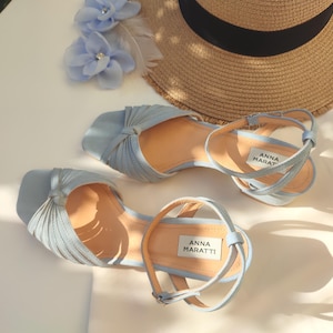 Może przedstawiać: Jasnoniebieskie sandały z paskami na kostkę i węzłowym wzorem na palcach. Sandały mają jasnobrązową wkładkę, a widoczna jest nazwa marki "ANNA MARATTI". Na zdjęciu widoczny jest r&oacute;wnież słomkowy kapelusz i niebieskie kwiatowe akcenty.