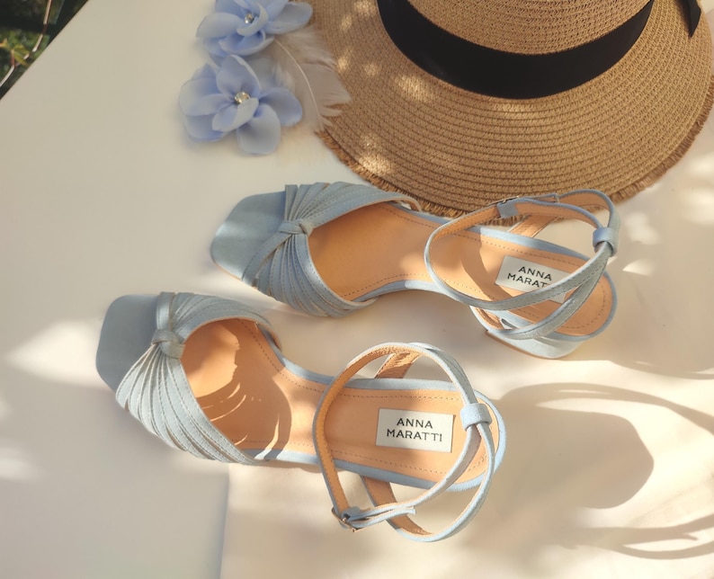 Może przedstawiać: Jasnoniebieskie sandały z paskami na kostkę i węzłowym wzorem na palcach. Sandały mają jasnobrązową wkładkę, a widoczna jest nazwa marki "ANNA MARATTI". Na zdjęciu widoczny jest r&oacute;wnież słomkowy kapelusz i niebieskie kwiatowe akcenty.