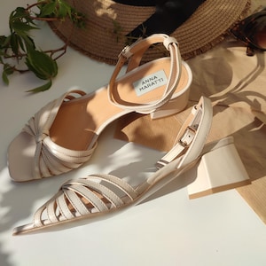 May include: A pair of beige leather sandals with a block heel. The sandals have a strappy design and an ankle strap with a buckle. The brand name "ANNA MARATTI" is visible on the insole. A straw hat and sunglasses are in the background.