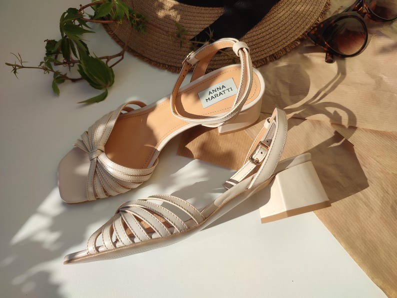 May include: A pair of beige leather sandals with a block heel. The sandals have a strappy design and an ankle strap with a buckle. The brand name "ANNA MARATTI" is visible on the insole. A straw hat and sunglasses are in the background.