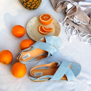 Pode incluir: Uma cena de ver&atilde;o com sand&aacute;lias azul claro, laranjas, um abacaxi e uma toalha bege. As sand&aacute;lias, com a etiqueta "ANNA MARATTI", t&ecirc;m palmilhas laranja. A cena est&aacute; sobre uma superf&iacute;cie branca.