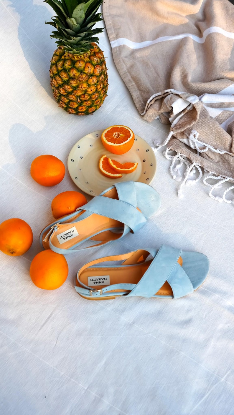 Pode incluir: Uma cena de ver&atilde;o com sand&aacute;lias azul claro, laranjas, um abacaxi e uma toalha bege. As sand&aacute;lias, com a etiqueta "ANNA MARATTI", t&ecirc;m palmilhas laranja. A cena est&aacute; sobre uma superf&iacute;cie branca.