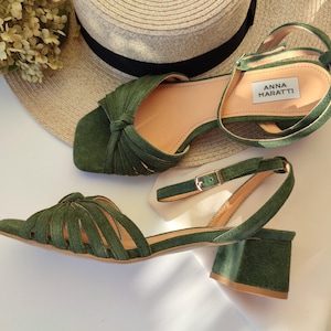 Może przedstawiać: Zielone sandały w kolorze oliwkowym z klockowym obcasem i paskiem na kostkę. Sandały mają wiązany wz&oacute;r na pasku na palce i zapięcie na klamrę. Nazwa marki "ANNA MARATTI" jest widoczna na wkładce. Słomkowy kapelusz i suszone kwiaty są w tle.