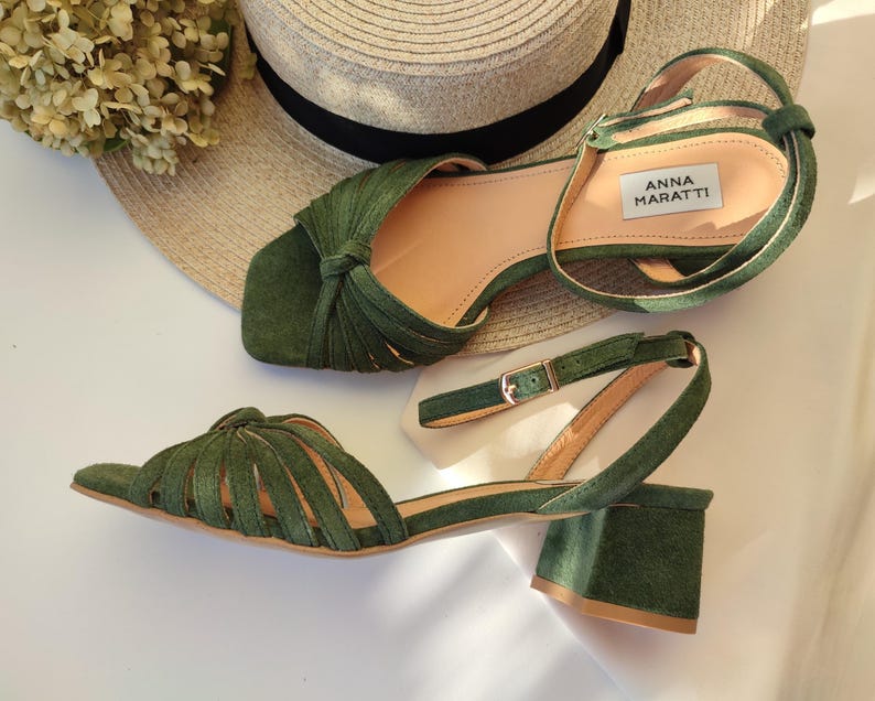 Może przedstawiać: Zielone sandały w kolorze oliwkowym z klockowym obcasem i paskiem na kostkę. Sandały mają wiązany wz&oacute;r na pasku na palce i zapięcie na klamrę. Nazwa marki "ANNA MARATTI" jest widoczna na wkładce. Słomkowy kapelusz i suszone kwiaty są w tle.