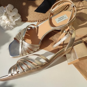 May include: Gold-colored sandals with a square toe and a small block heel. The sandals have multiple straps across the foot and ankle straps with buckles. The brand name "ANNA MARATTI" is visible on a label on one of the sandals. A straw hat and white flowers are in the background.