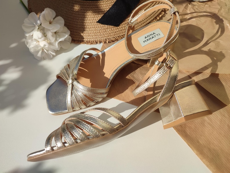 May include: Gold-colored sandals with a square toe and a small block heel. The sandals have multiple straps across the foot and ankle straps with buckles. The brand name "ANNA MARATTI" is visible on a label on one of the sandals. A straw hat and white flowers are in the background.