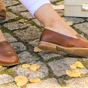 Pu&ograve; includere: Mocassini in pelle marrone con tacco basso. Le scarpe sono indossate con pantaloni bianchi e poggiano su una superficie di ciottoli con foglie gialle cadute. Una borsa color crema &egrave; sullo sfondo.