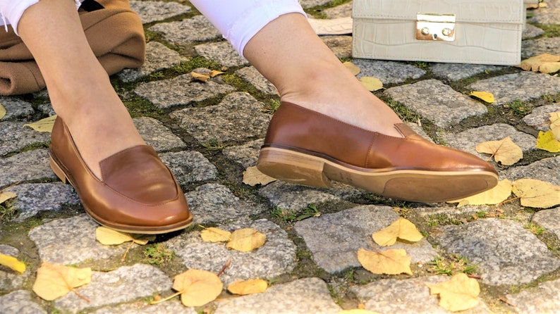 Pu&ograve; includere: Mocassini in pelle marrone con tacco basso. Le scarpe sono indossate con pantaloni bianchi e poggiano su una superficie di ciottoli con foglie gialle cadute. Una borsa color crema &egrave; sullo sfondo.