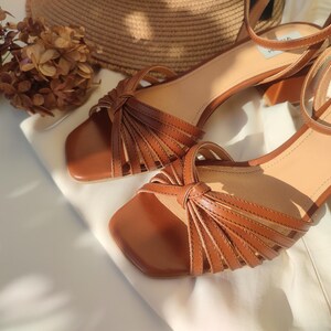 Puede incluir: Sandalias de cuero marr&oacute;n con punta cuadrada y correa en el tobillo. Las sandalias tienen un dise&ntilde;o anudado en la parte delantera y un tac&oacute;n peque&ntilde;o. El nombre de la marca "ANNA MARATTI" es visible en una sandalia. Las sandalias se muestran sobre una superficie blanca.