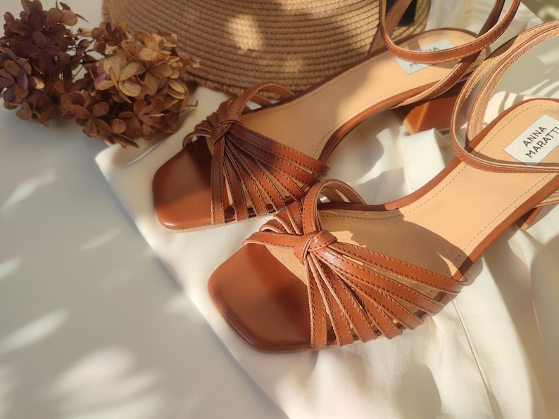 Puede incluir: Sandalias de cuero marr&oacute;n con punta cuadrada y correa en el tobillo. Las sandalias tienen un dise&ntilde;o anudado en la parte delantera y un tac&oacute;n peque&ntilde;o. El nombre de la marca "ANNA MARATTI" es visible en una sandalia. Las sandalias se muestran sobre una superficie blanca.