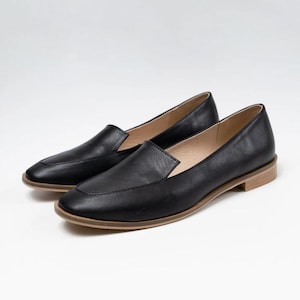 Pu&ograve; includere: Un paio di mocassini in pelle nera con punta quadrata e tacco basso color chiaro. Le scarpe sono esposte su uno sfondo bianco, mostrando il loro design elegante e l'estetica minimalista.