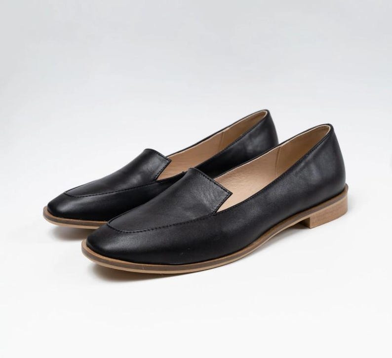 Pu&ograve; includere: Un paio di mocassini in pelle nera con punta quadrata e tacco basso color chiaro. Le scarpe sono esposte su uno sfondo bianco, mostrando il loro design elegante e l'estetica minimalista.