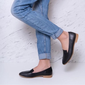 Pu&ograve; includere: Mocassini in pelle nera con suola marrone chiaro. Le scarpe hanno un classico design slip-on e sono indossate con jeans blu. I mocassini sono un'opzione di calzatura versatile per occasioni casual o semi-formali.