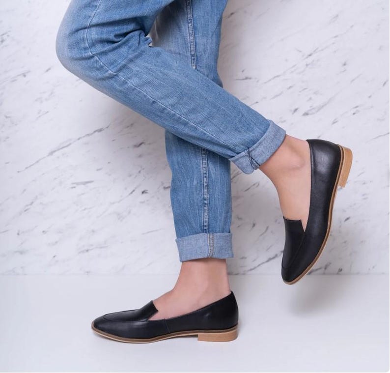 Pu&ograve; includere: Mocassini in pelle nera con suola marrone chiaro. Le scarpe hanno un classico design slip-on e sono indossate con jeans blu. I mocassini sono un'opzione di calzatura versatile per occasioni casual o semi-formali.