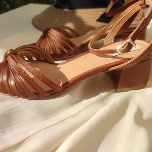 Puede incluir: Sandalias de cuero marr&oacute;n con punta cuadrada y correa en el tobillo. Las sandalias tienen tac&oacute;n de bloque y un dise&ntilde;o tejido en la parte delantera. Un sombrero de paja est&aacute; en el fondo.