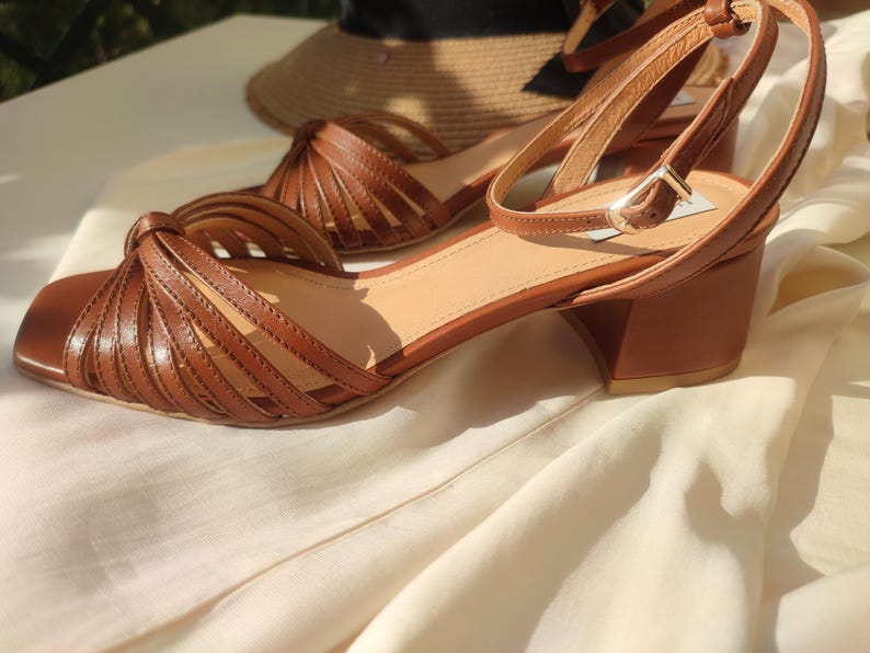 Puede incluir: Sandalias de cuero marr&oacute;n con punta cuadrada y correa en el tobillo. Las sandalias tienen tac&oacute;n de bloque y un dise&ntilde;o tejido en la parte delantera. Un sombrero de paja est&aacute; en el fondo.