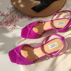 Może przedstawiać: Fuksjowe sandały z kwadratowym noskiem i paskiem na kostkę. Sandały mają niski obcas. Nazwa marki "ANNA MARATTI" jest nadrukowana na wkładce. Słomkowy kapelusz i r&oacute;żowe kwiaty są w tle.