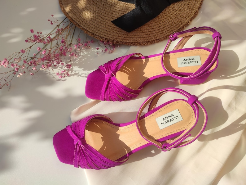 Może przedstawiać: Fuksjowe sandały z kwadratowym noskiem i paskiem na kostkę. Sandały mają niski obcas. Nazwa marki "ANNA MARATTI" jest nadrukowana na wkładce. Słomkowy kapelusz i r&oacute;żowe kwiaty są w tle.