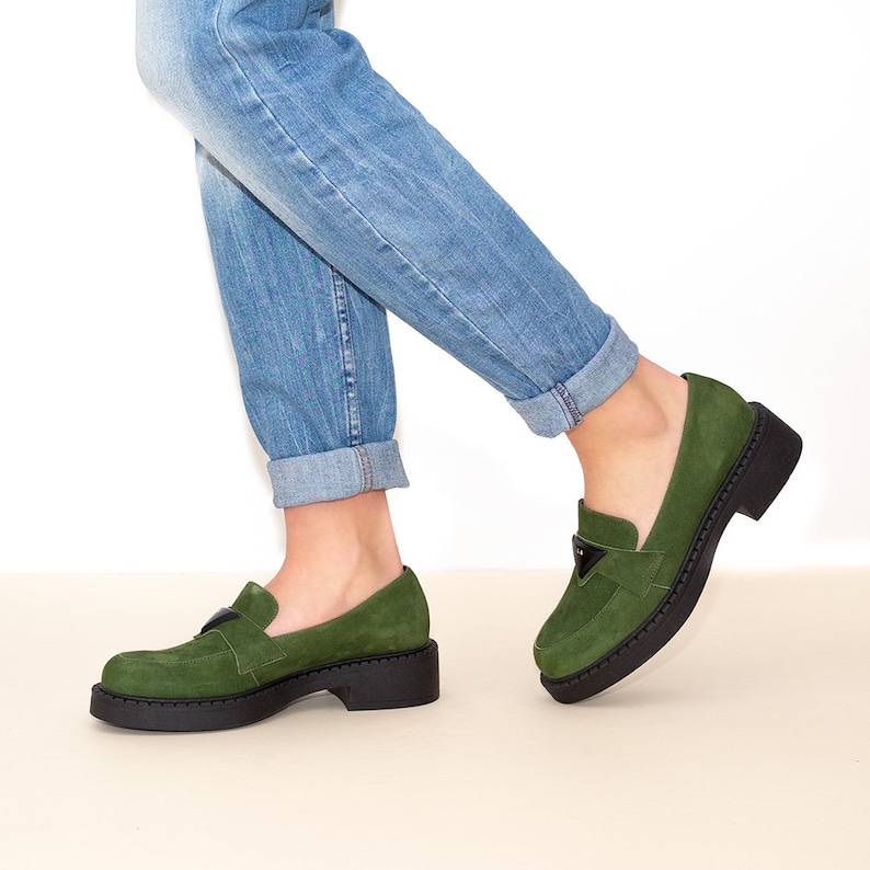May include: Green suede loafers with a black platform sole and a gold buckle detail. The loafers are worn with blue jeans.