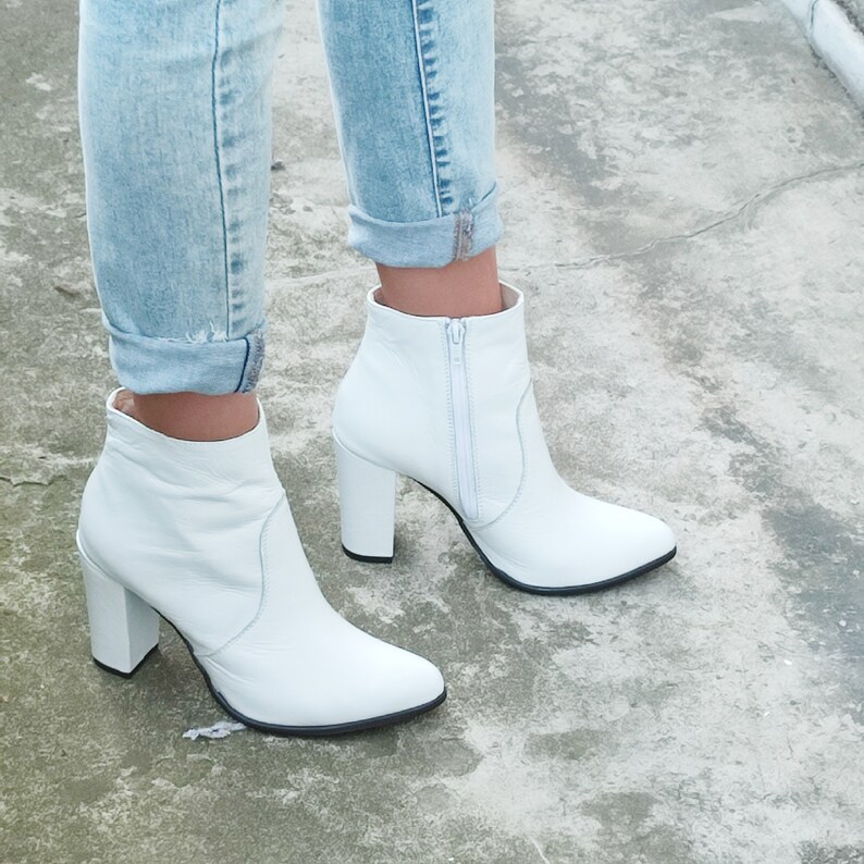 May include: White leather ankle boots with a block heel and a zipper up the back. The boots are worn with light blue jeans.