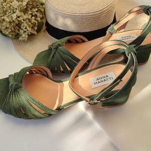 Może przedstawiać: Zielone sandały zamszowe z wiązanym wzorem i paskami na kostkę. Sandały mają niski obcas w kształcie klocka i kwadratowy nosek. Marka "ANNA MARATTI" jest widoczna na wkładce. Słomkowy kapelusz i suszone kwiaty są w tle.