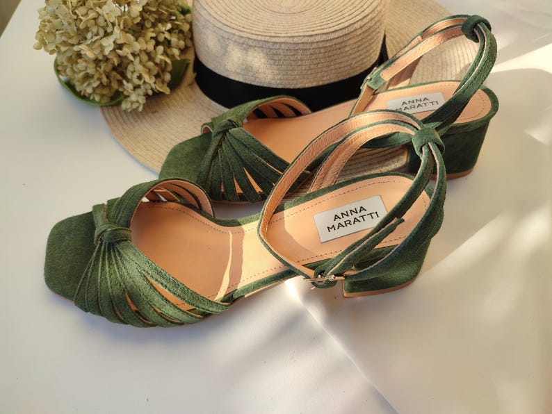 Może przedstawiać: Zielone sandały zamszowe z wiązanym wzorem i paskami na kostkę. Sandały mają niski obcas w kształcie klocka i kwadratowy nosek. Marka "ANNA MARATTI" jest widoczna na wkładce. Słomkowy kapelusz i suszone kwiaty są w tle.