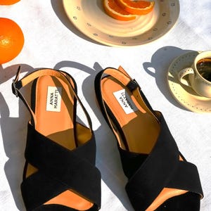 Pode incluir: Sand&aacute;lias pretas de camur&ccedil;a com tiras cruzadas, com a marca "ANNA MARATTI" na palmilha. A imagem inclui laranjas, um abacaxi e uma x&iacute;cara de caf&eacute;, sugerindo um tema de estilo de vida.