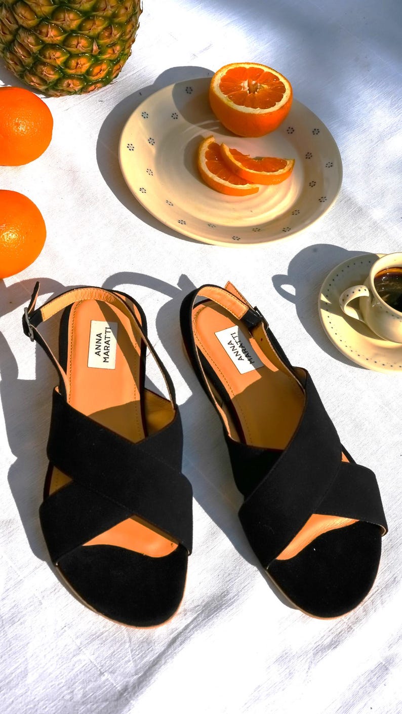 Pode incluir: Sand&aacute;lias pretas de camur&ccedil;a com tiras cruzadas, com a marca "ANNA MARATTI" na palmilha. A imagem inclui laranjas, um abacaxi e uma x&iacute;cara de caf&eacute;, sugerindo um tema de estilo de vida.