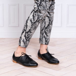 May include: Black leather lace-up shoes with a rounded toe. The shoes are worn by a person wearing black and white patterned pants.