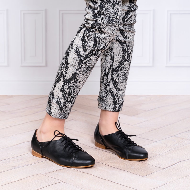 May include: Black leather lace-up shoes with a rounded toe. The shoes are worn by a person wearing black and white patterned pants.