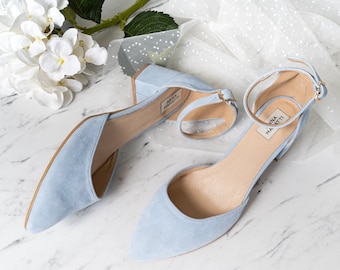 Puede incluir: Un par de zapatos de tacón de ante azul claro con correas de tobillo. Los zapatos tienen una punta puntiaguda y un tacón de bloque. El nombre de la marca "Anna Maratti" es visible en la plantilla de cada zapato.