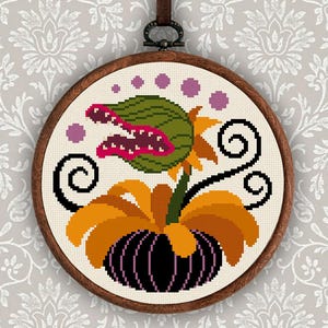 May include: A cross-stitch embroidery in a wooden hoop features a green, red, and purple plant with a large mouth. The plant has orange and yellow petals and a black base. The embroidery is suspended by a brown leather strap.