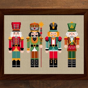 May include: A framed cross-stitch featuring four colorful nutcracker figures in red, green, and blue uniforms. The artwork is displayed on a neutral background, surrounded by Christmas decorations, including wrapped gifts, pine cones, and ornaments.
