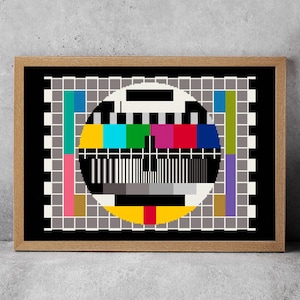 May include: A framed artwork featuring a vintage TV test pattern. The colorful circular pattern is set against a black background and a grid of white squares. The frame is made of light brown wood. A black vase with dried flowers is to the right.